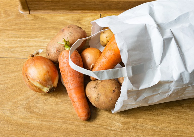 Foto de batata, cenoura e cebola. Metade dos alimentos está do lado de fora de uma sacola de compras. Os alimentos na sacola estão por cima de uma mesa.